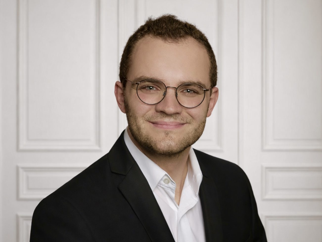 Ein junger Mann mit Brille, lächelnd in formeller Kleidung vor einer neutralen Wand.