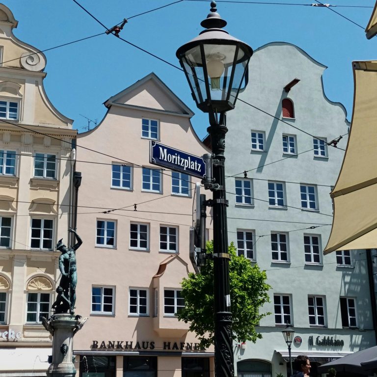 Farbenfrohe Gebäude an einem Platz mit Straßenlaterne und grüner Pflanze im Vordergrund.
